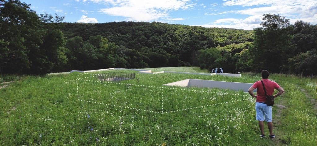 Ein Mann steht auf einer Wiese neben den weißen Betonwänden eines Erdhauses, die aus dem Gras ragen.