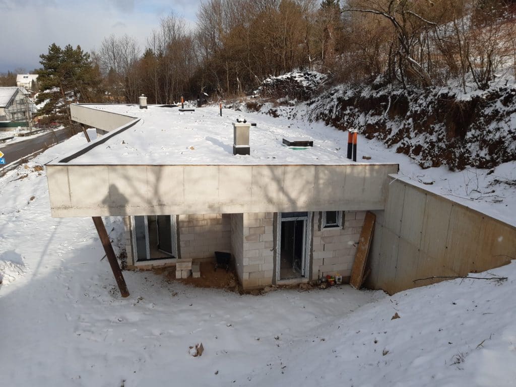 Schneebedecktes Betondach eines im Bau befindlichen Erdhauses, darunter Mauerwerk mit noch unverglasten Öffnungen