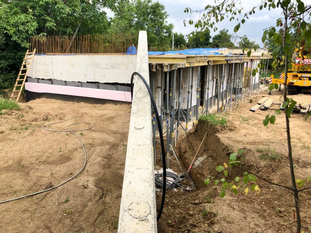 Baustelle eines Erdhauses: Betonwand in der Schalung, daneben fertiger Abschnitt mit Dichtungsbahn und rosa Dämmplatten, Bewehrung ragt heraus, Kran im Hintergrund.