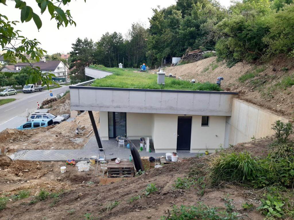 Erdhaus im Bau, Ansicht von unten: helle Putzfassade, Terrasse, schräge Betonwand an aufgeschütteter Erde, begrüntes Dach.