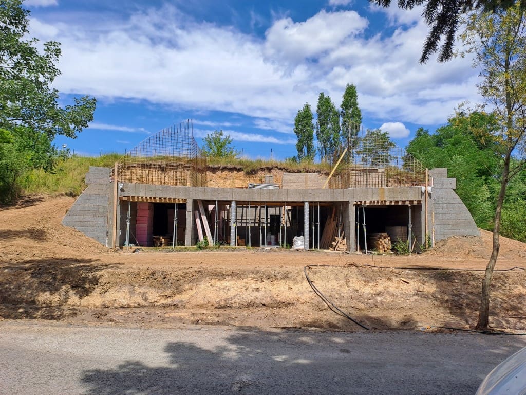 Rozestavěný podzemní dům, betonová konstrukce zapuštěná do svahu, vzadu už zatravněný násep nad stavbou.