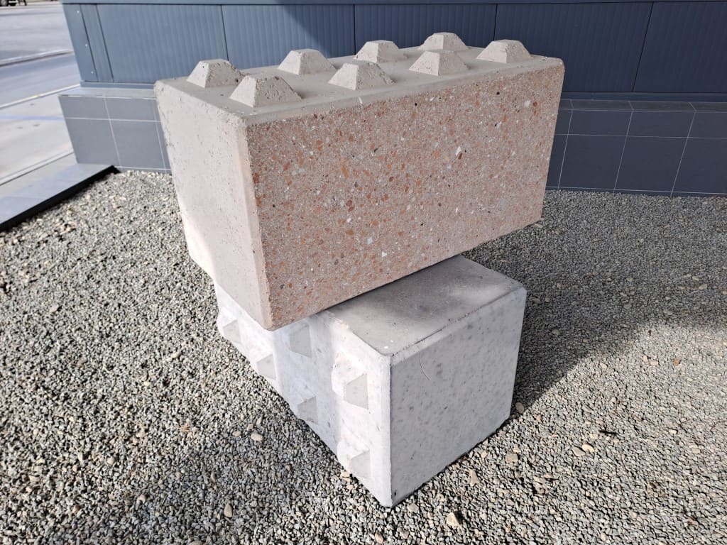 Dva prefabrikované betonové bloky s propojovacími čepy uložené na štěrku před šedým opláštěním