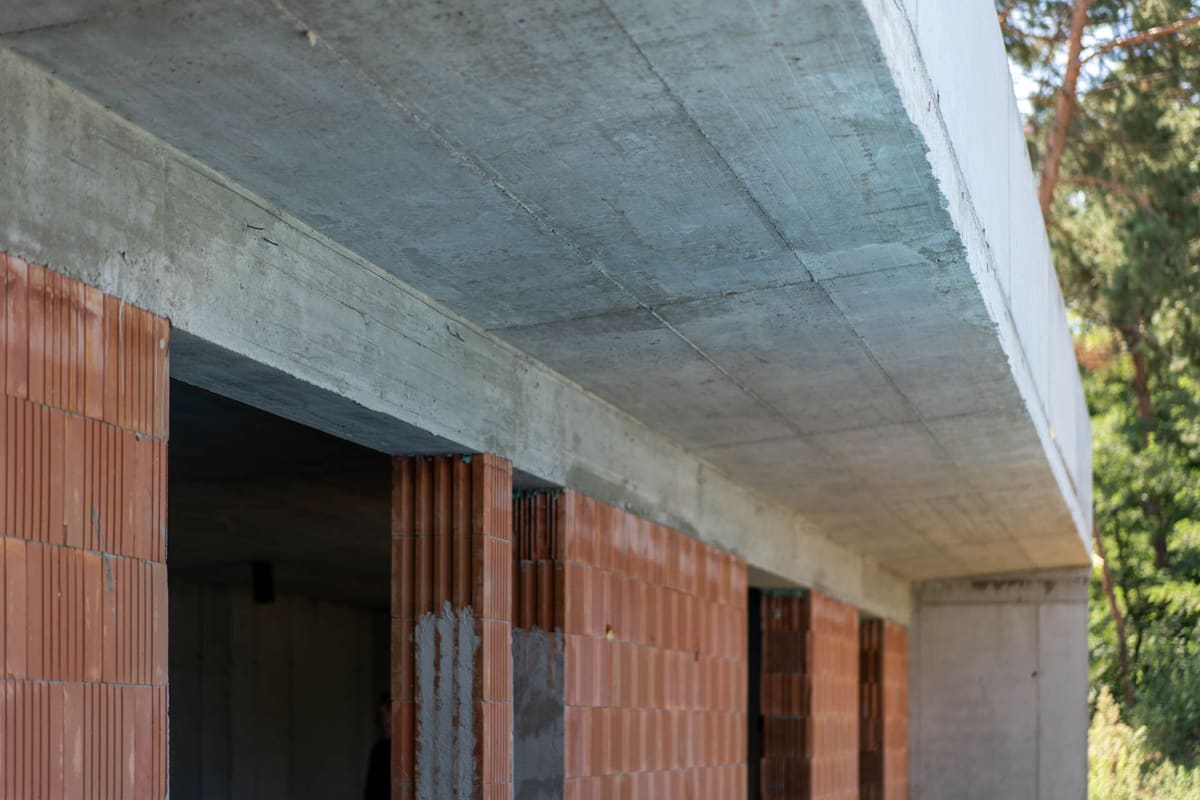 Baustelle mit auskragender Betonplatte über einer Wand aus gelochten Ziegeln, Bewehrungsstäbe ragen hervor.