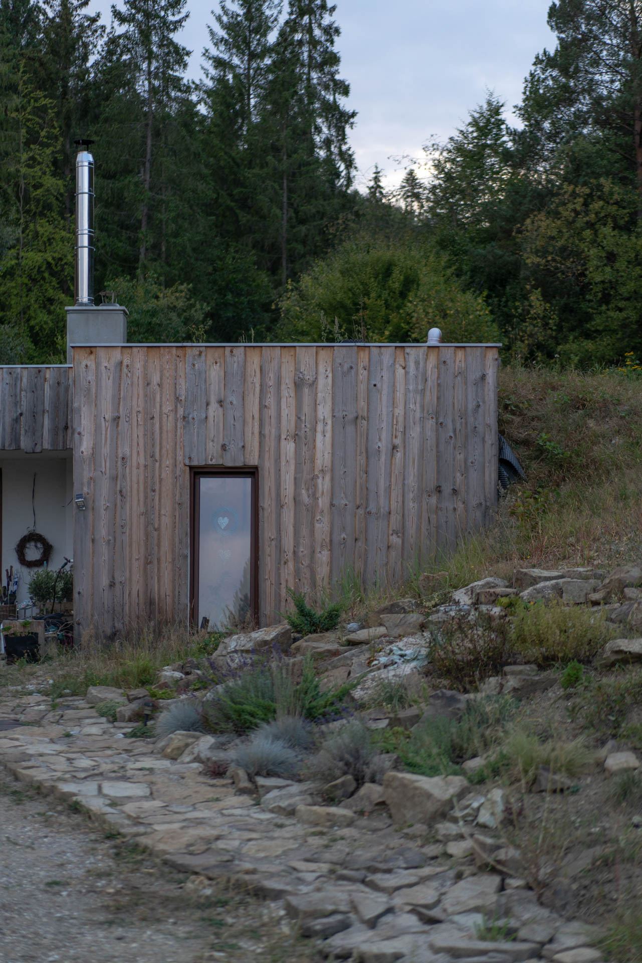 Parete di testata in legno della casa interrata Top Home, con porta vetrata e terra e erba che salgono verso il bosco.