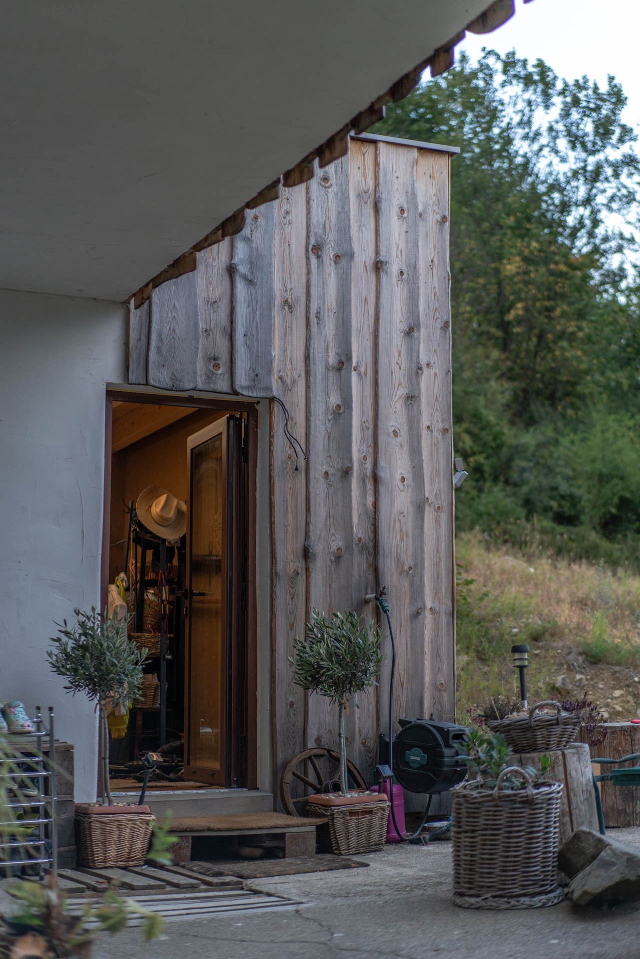 Ingresso rivestito in legno della casa interrata Top Home, terrazza con ulivi in vaso e ruota di carro