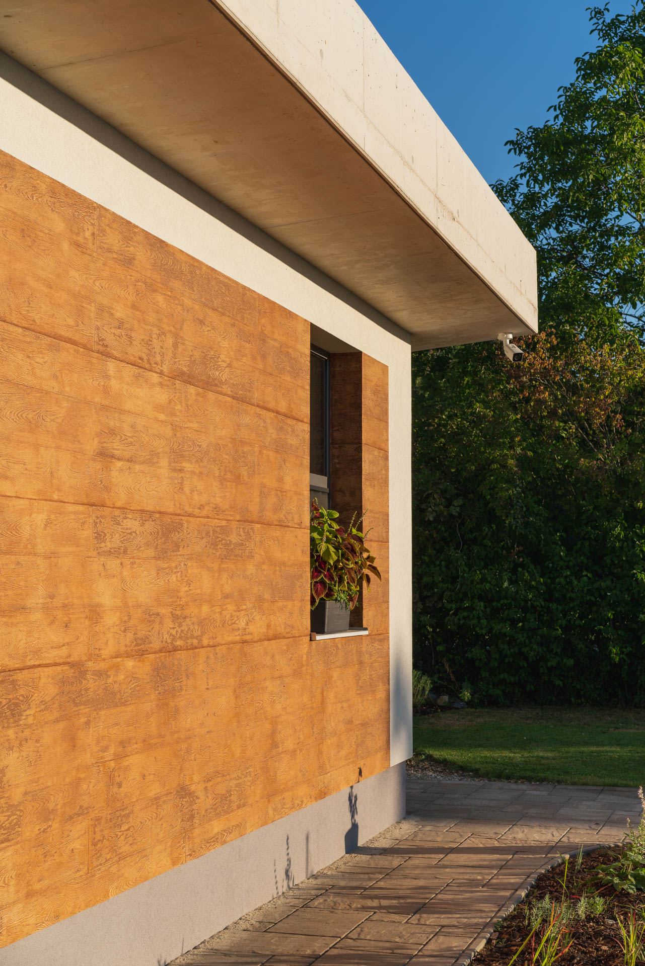 Wood-panelled corner of an earth-sheltered house in Nove Mesto nad Vahom, camera under the concrete roof overhang, window with plants.