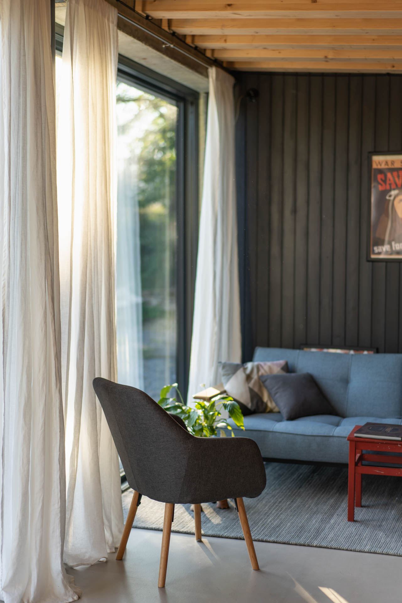 Living room of an earth-sheltered house in Banska Stiavnica, sheer curtains at a glazed door, blue sofa, dark wood wall