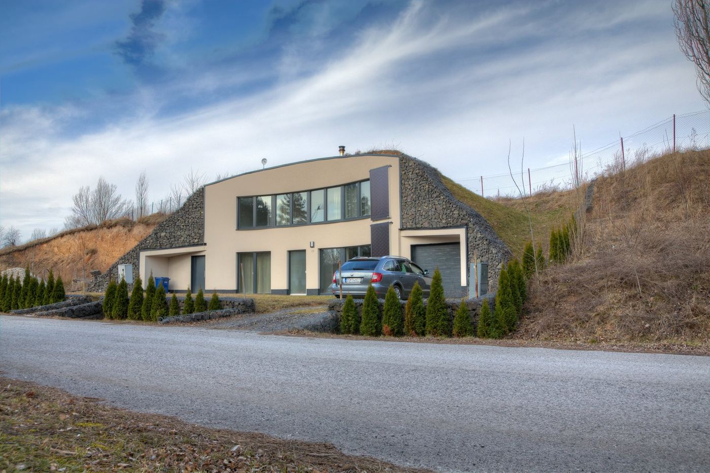 Earth-sheltered house in Rohoznik with cream front wall between grassy gabion-walled mounds and a parked car