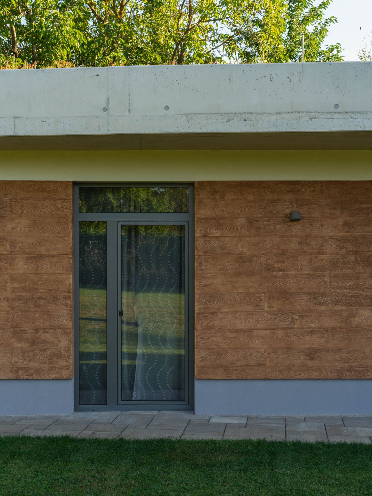 Wood-panelled entrance of an earth-sheltered house in Nove Mesto nad Vahom, concrete roof edge with grass and trees above.