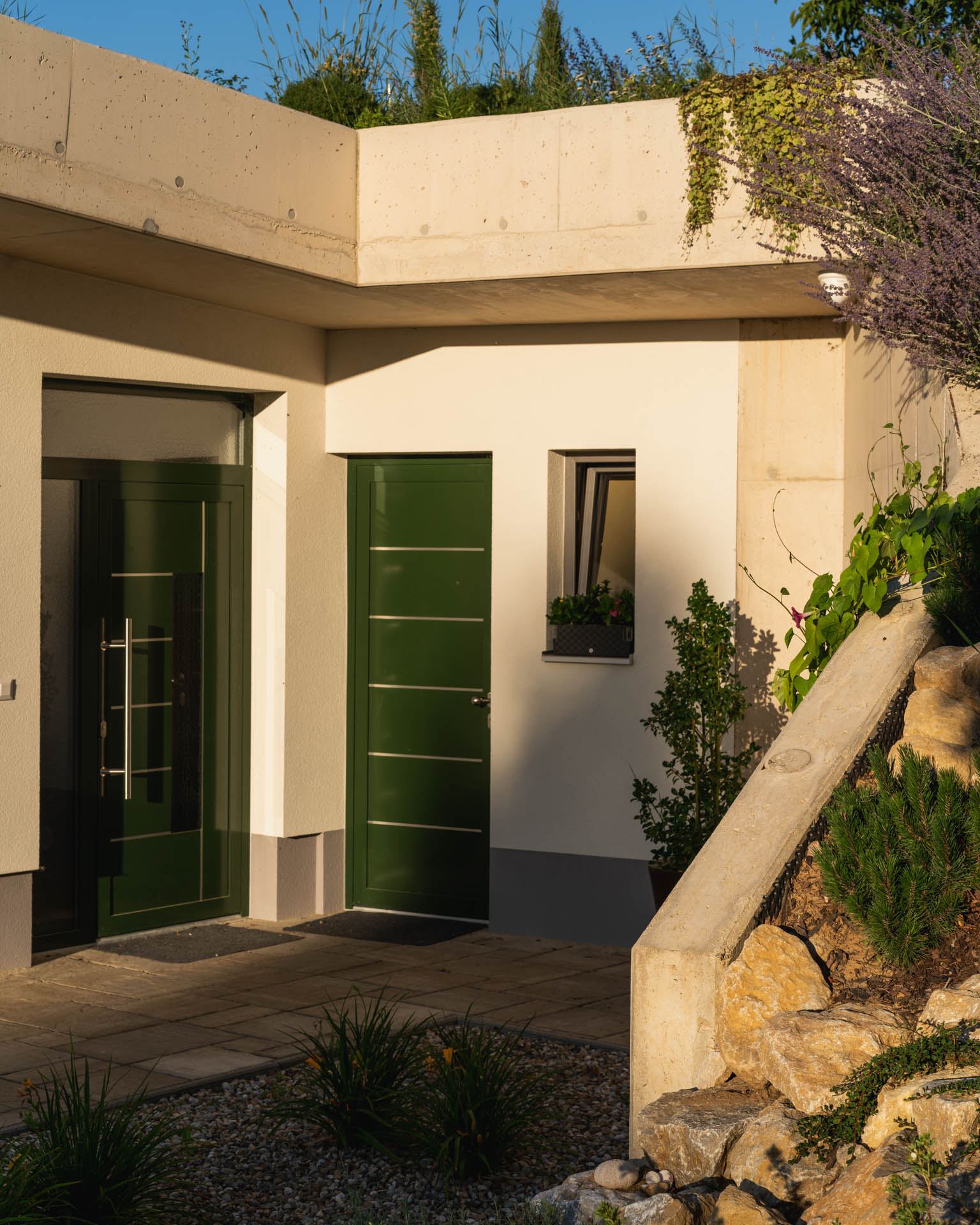Entrance corner of an earth-sheltered house in Nove Mesto nad Vahom, two green doors, grasses on the roof edge and a planted rocky slope beside a terrace.