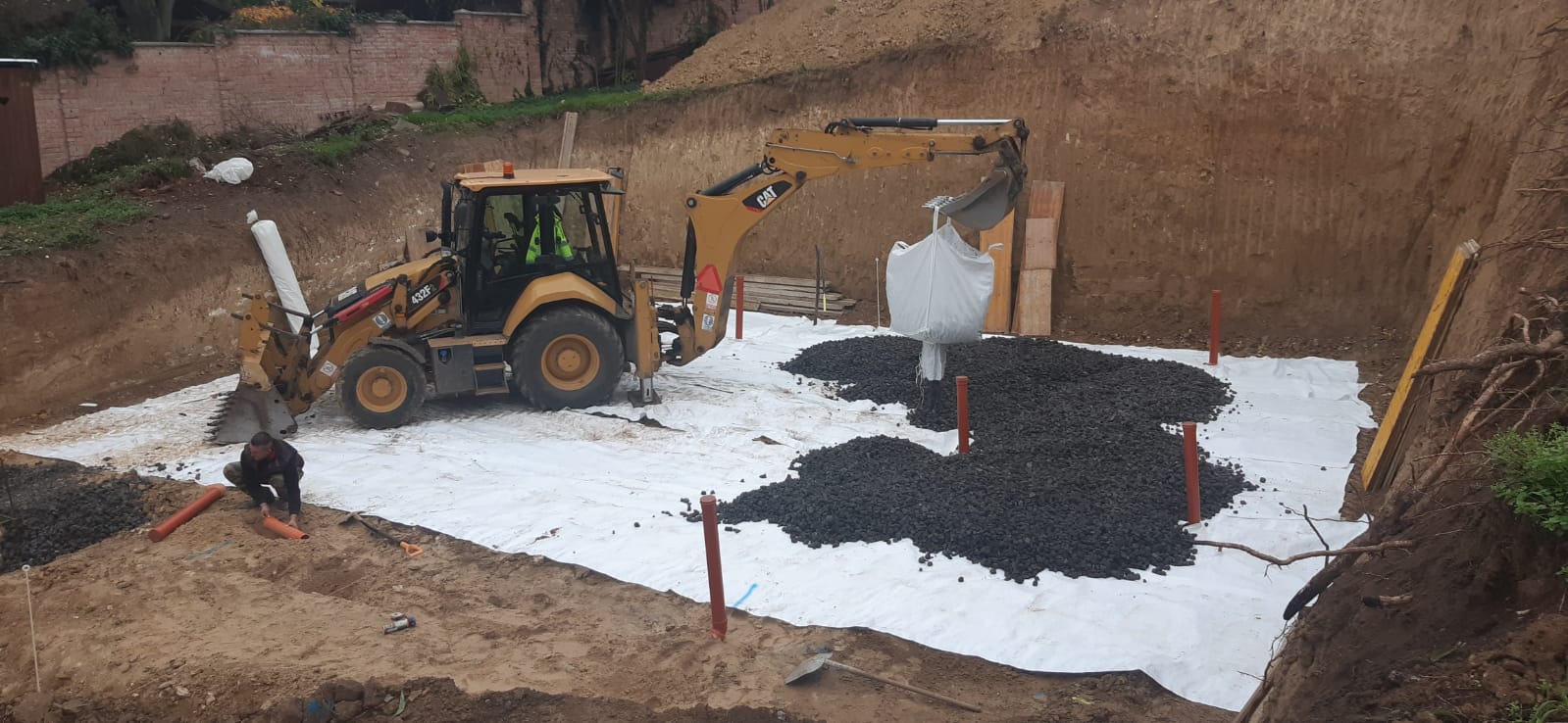 Scavo di fondazione della casa interrata di Trnava con geotessuto, cumuli di ghiaia, escavatore e operaio con tubo arancione.