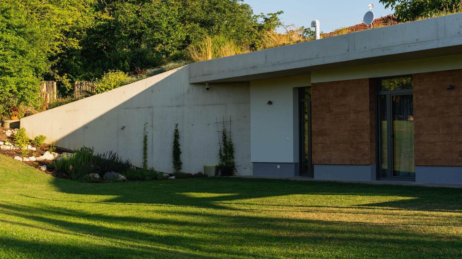 Casa interrata con parete di cemento inclinata e tetto verde, accanto porte a vetri e pannelli marrone ruggine
