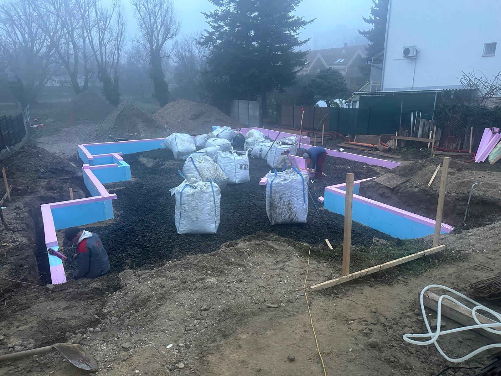 Foundation trench with blue and pink insulation panels for an earth-sheltered house study in Bratislava, workers at the edges