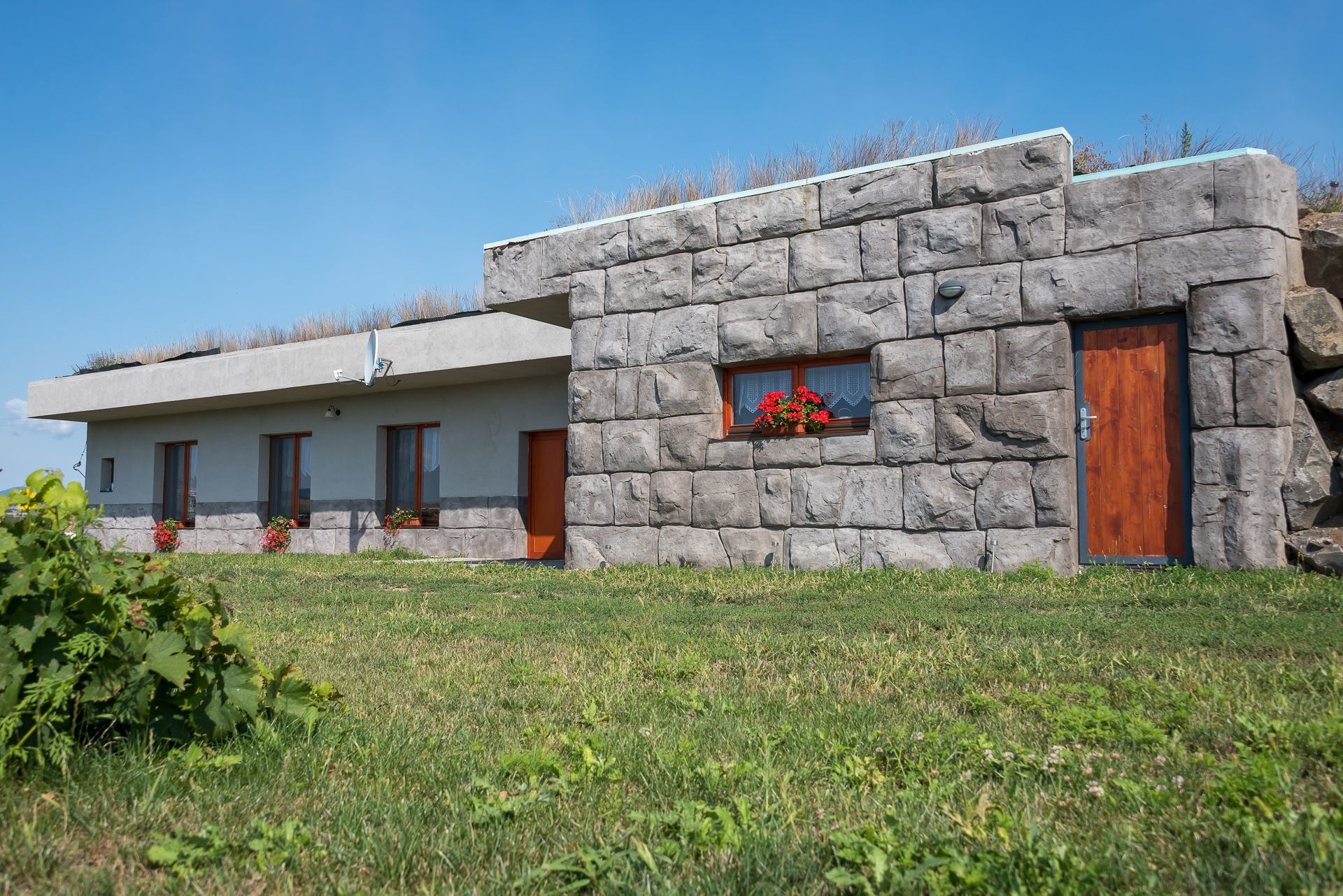 Earth-sheltered house Zemplin with stone-clad wall, geraniums and grassy roof edge on a green slope