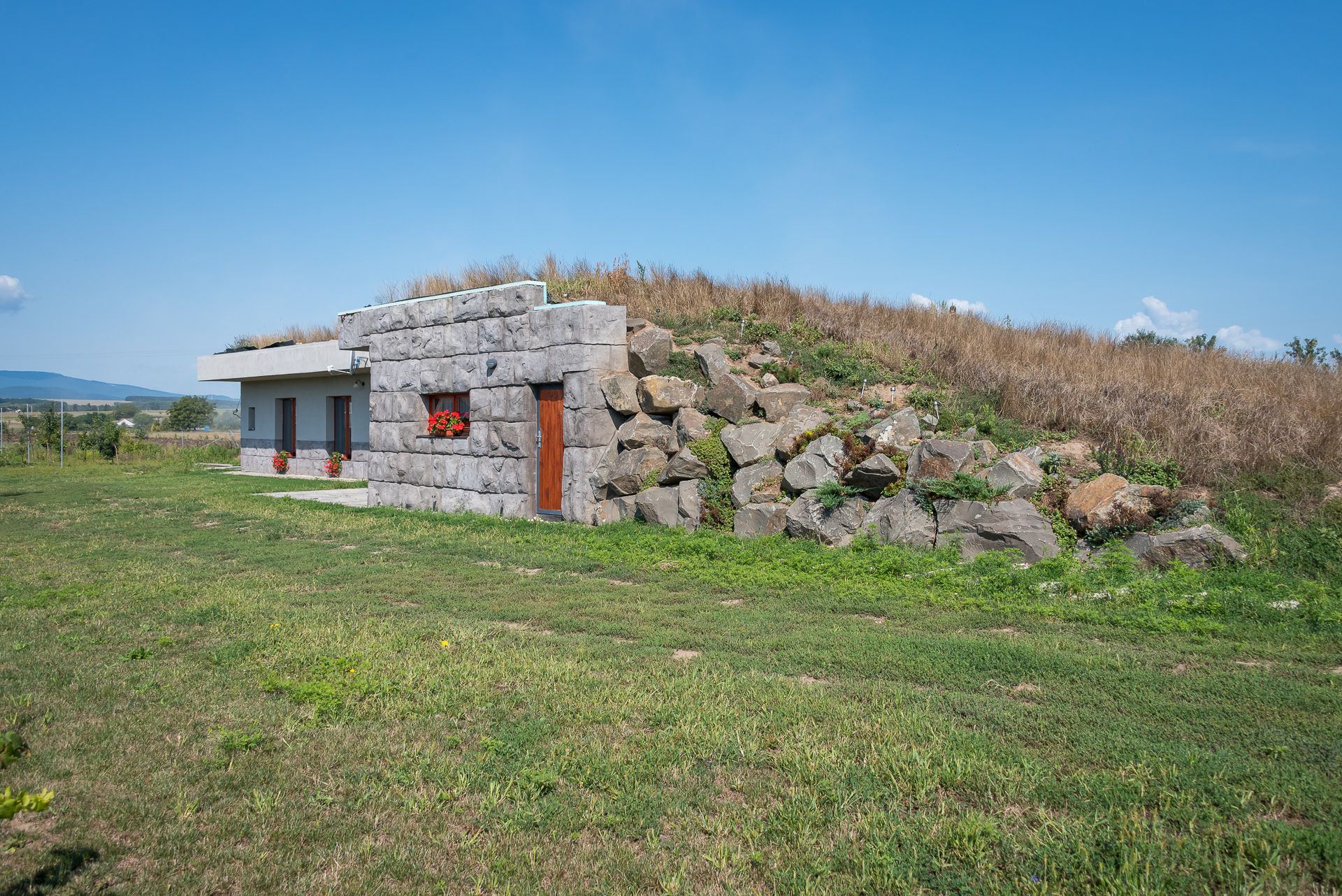 Earth-sheltered house Zemplin with stone-clad end wall, boulders on the mound and a grassy roof strip, grain field around it