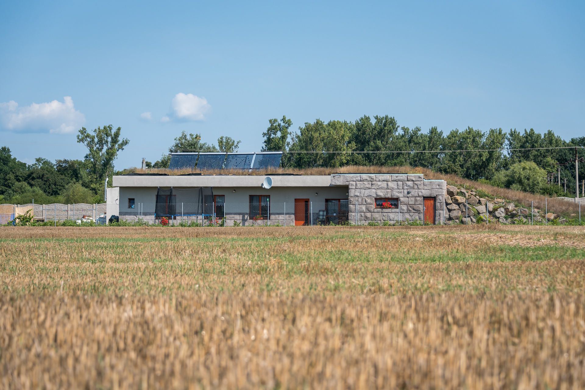 Earth-sheltered house Zemplin with grass roof, solar panels, orange window frames and a mound of boulders behind the stone gable