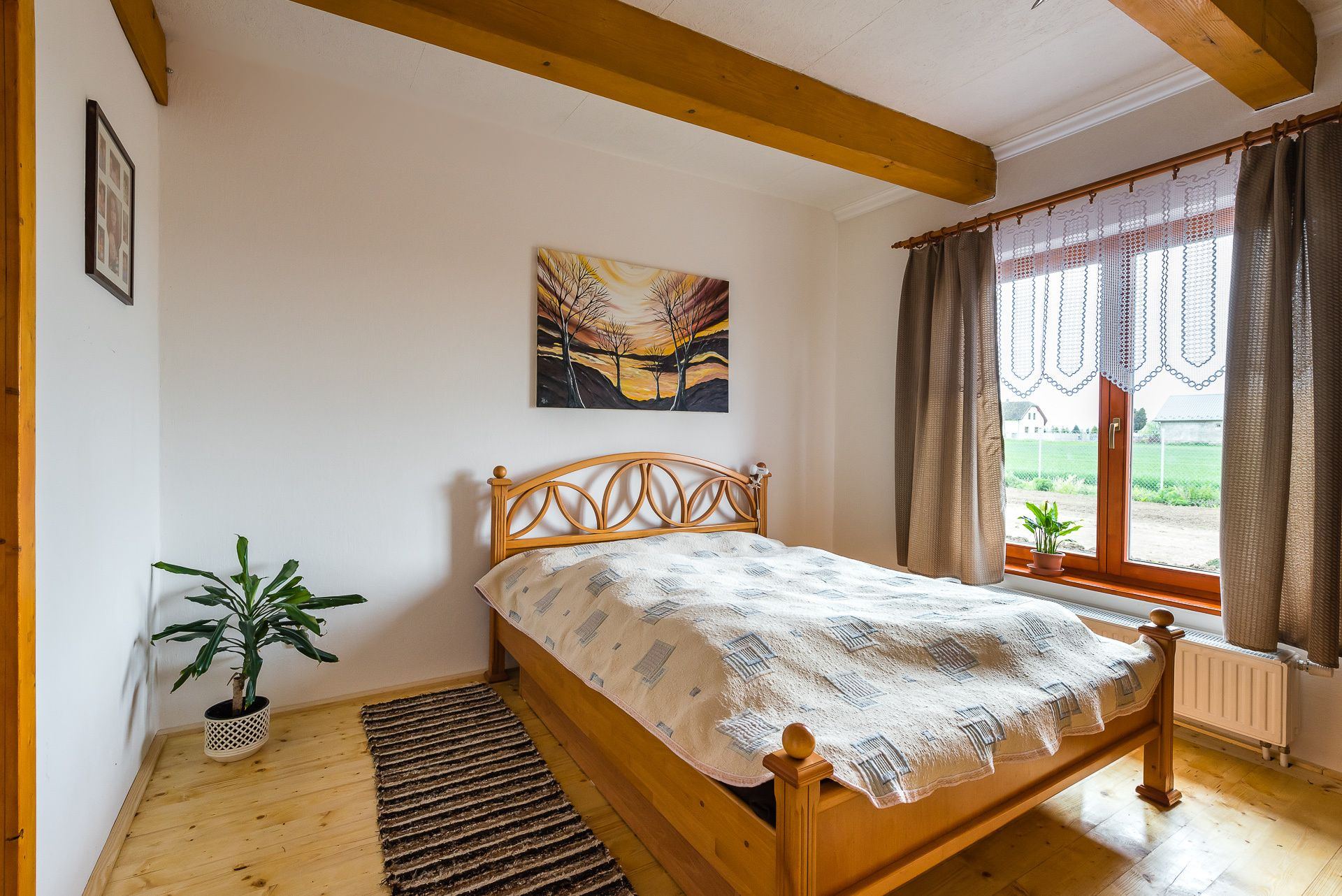 Bedroom in the Zemplin earth-sheltered house with wooden beams, a curved bed frame and a field view through wide curtained windows