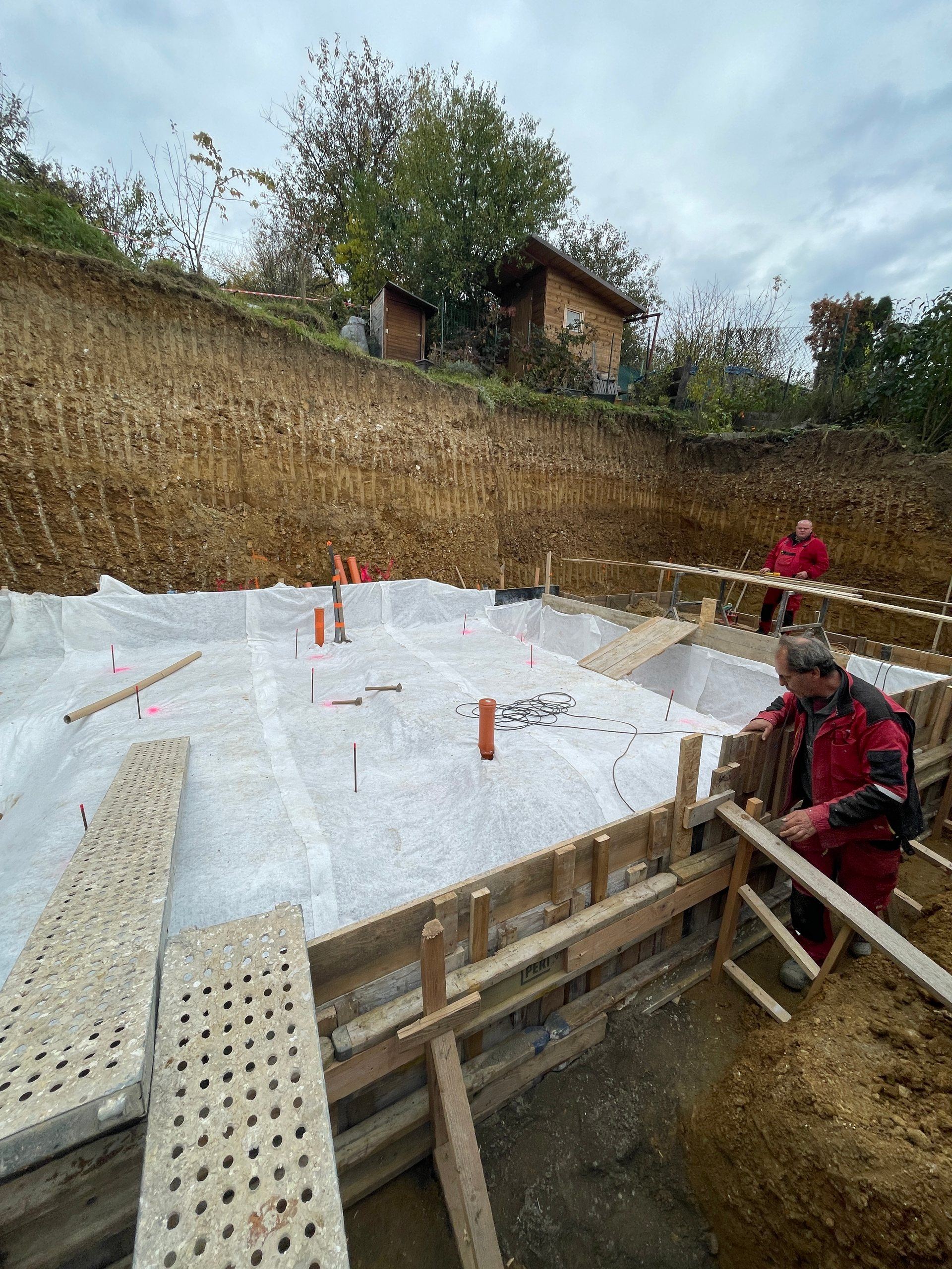 Baugrube mit PERI-Schalung und Geotextil für das Fundament des Erdhauses Terasa Košice, zwei Arbeiter im Einsatz.