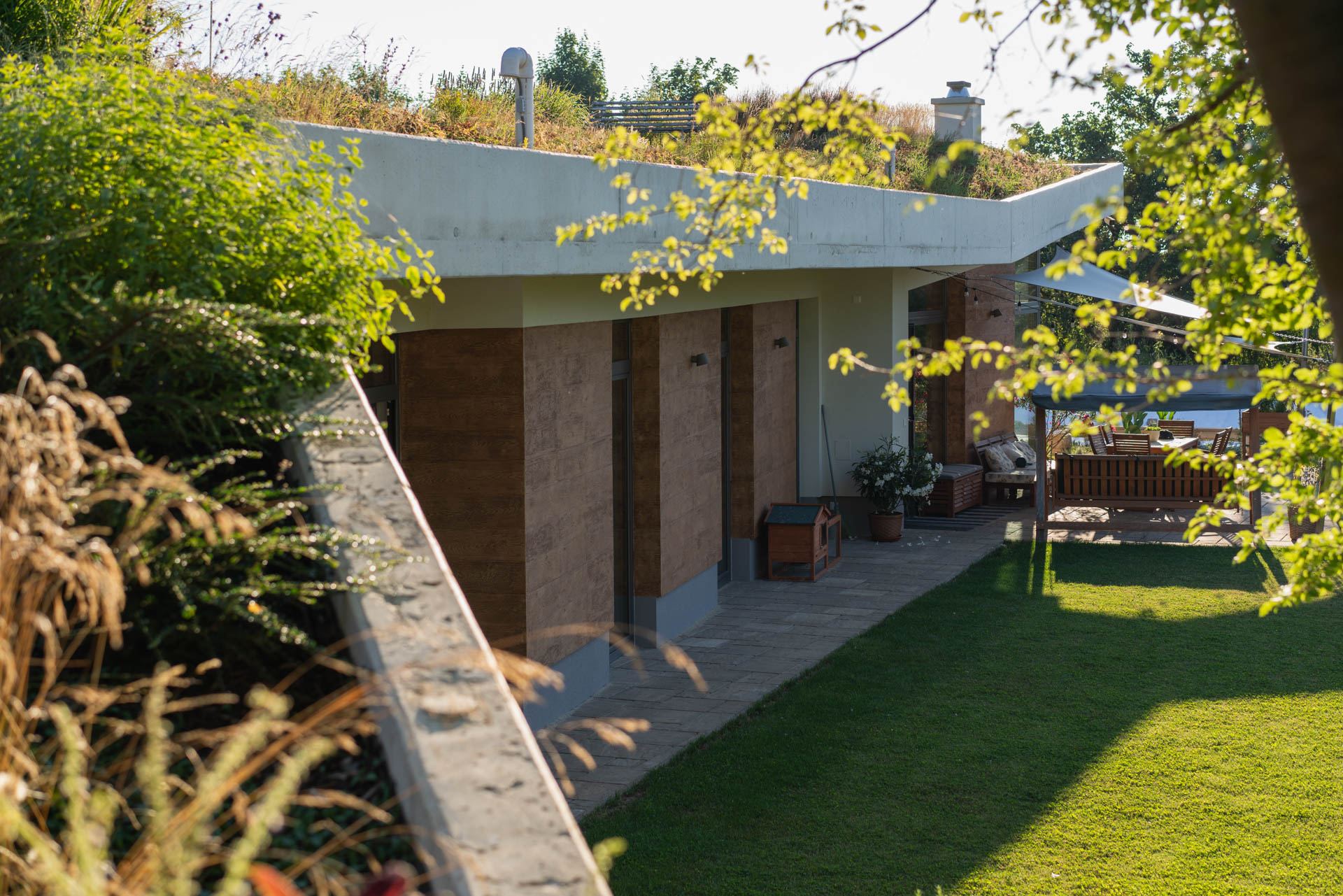 Earth-sheltered house in Nove Mesto nad Vahom, grass-covered roof over a shaded terrace, low concrete wall by the lawn.