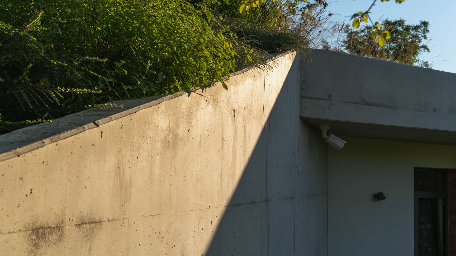 Bare concrete corner of an earth-sheltered house in Nove Mesto nad Vahom, roof edge planted with grasses, camera and light fixed below the overhang.