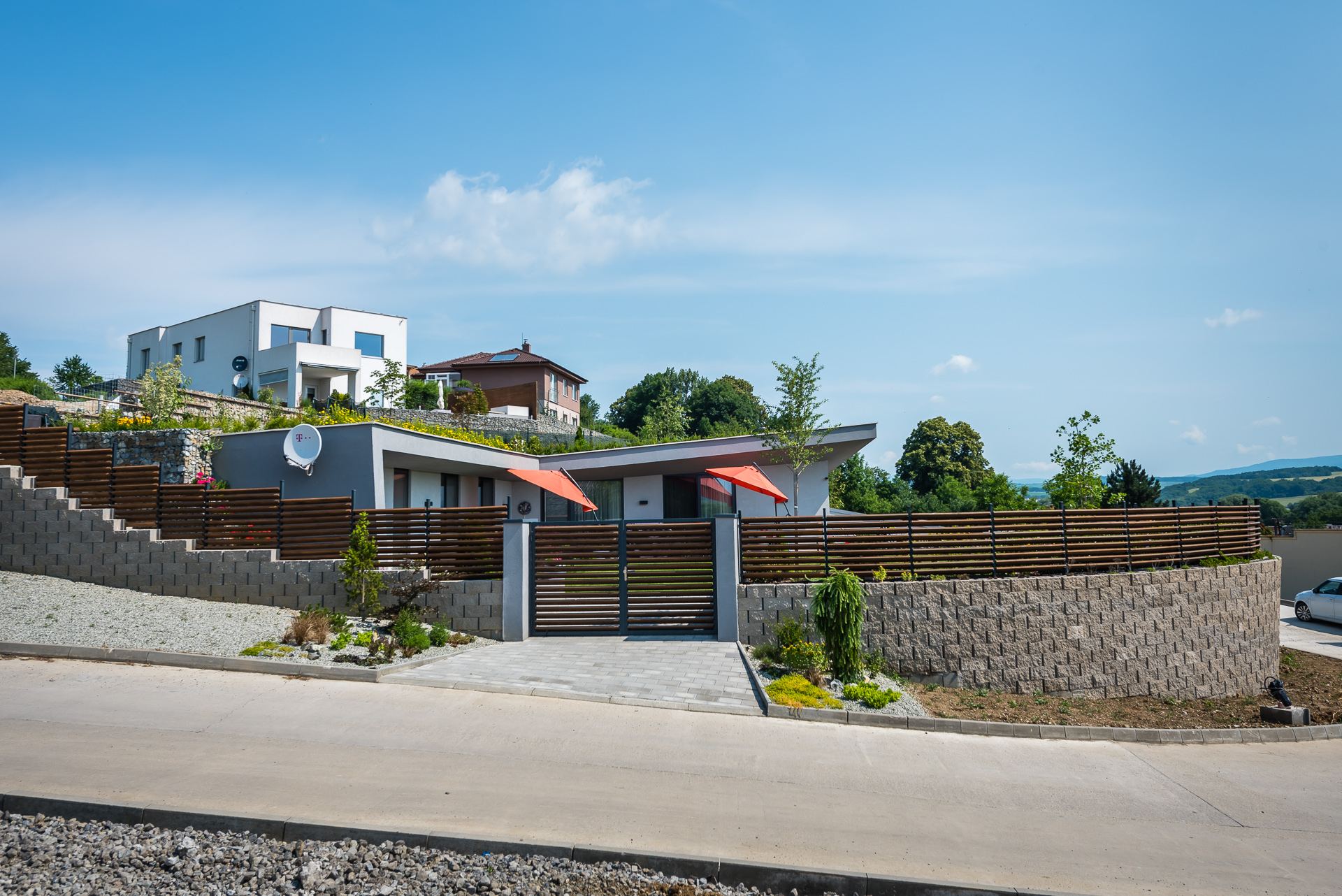 Earth-sheltered house Hoblina in Košice, grey facade behind a wooden fence, grass strip on the roof edge and stone terraces above.