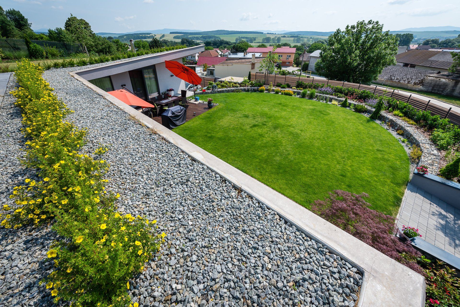 Aerial view of the earth-sheltered Hoblina house in Košice, gravel roof beds with shrubs above a terrace with red umbrellas and a round lawn.