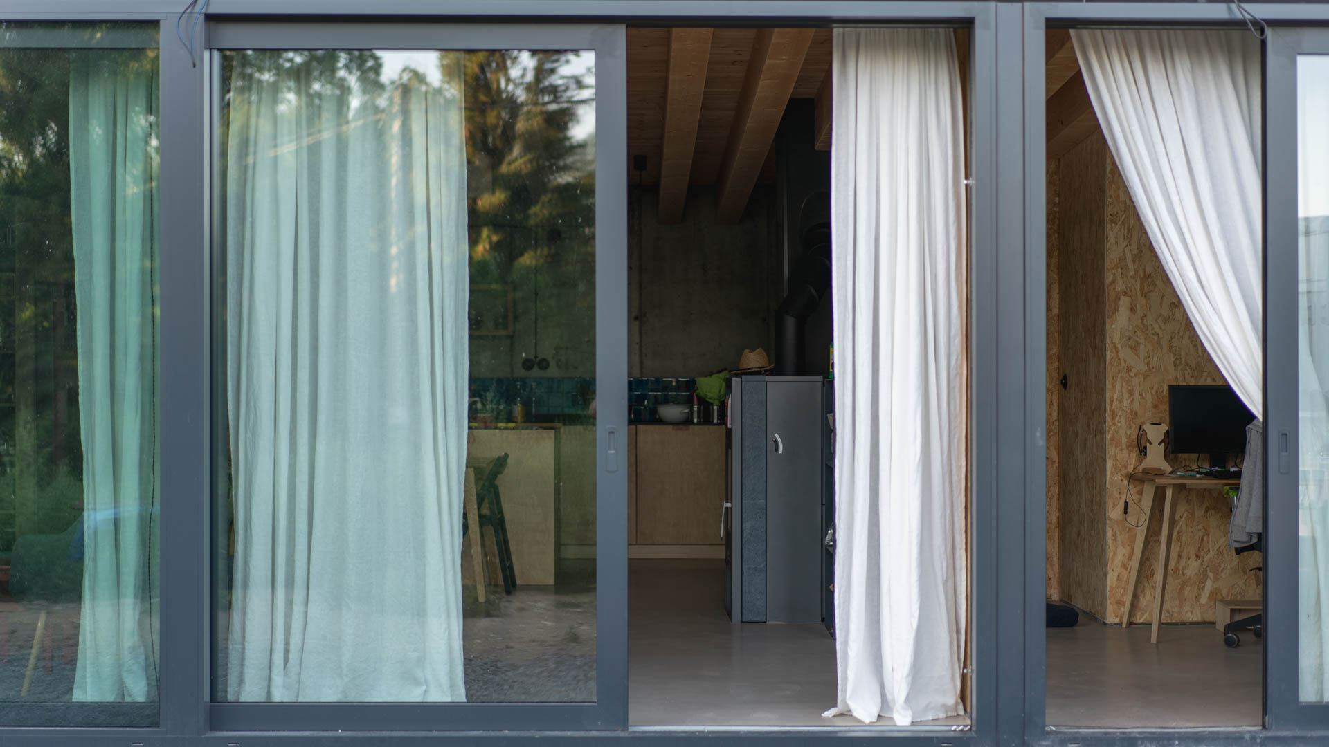 Glazed sliding facade of an earth-sheltered house in Banska Stiavnica, view into kitchen with wood ceiling beams and a study with OSB walls