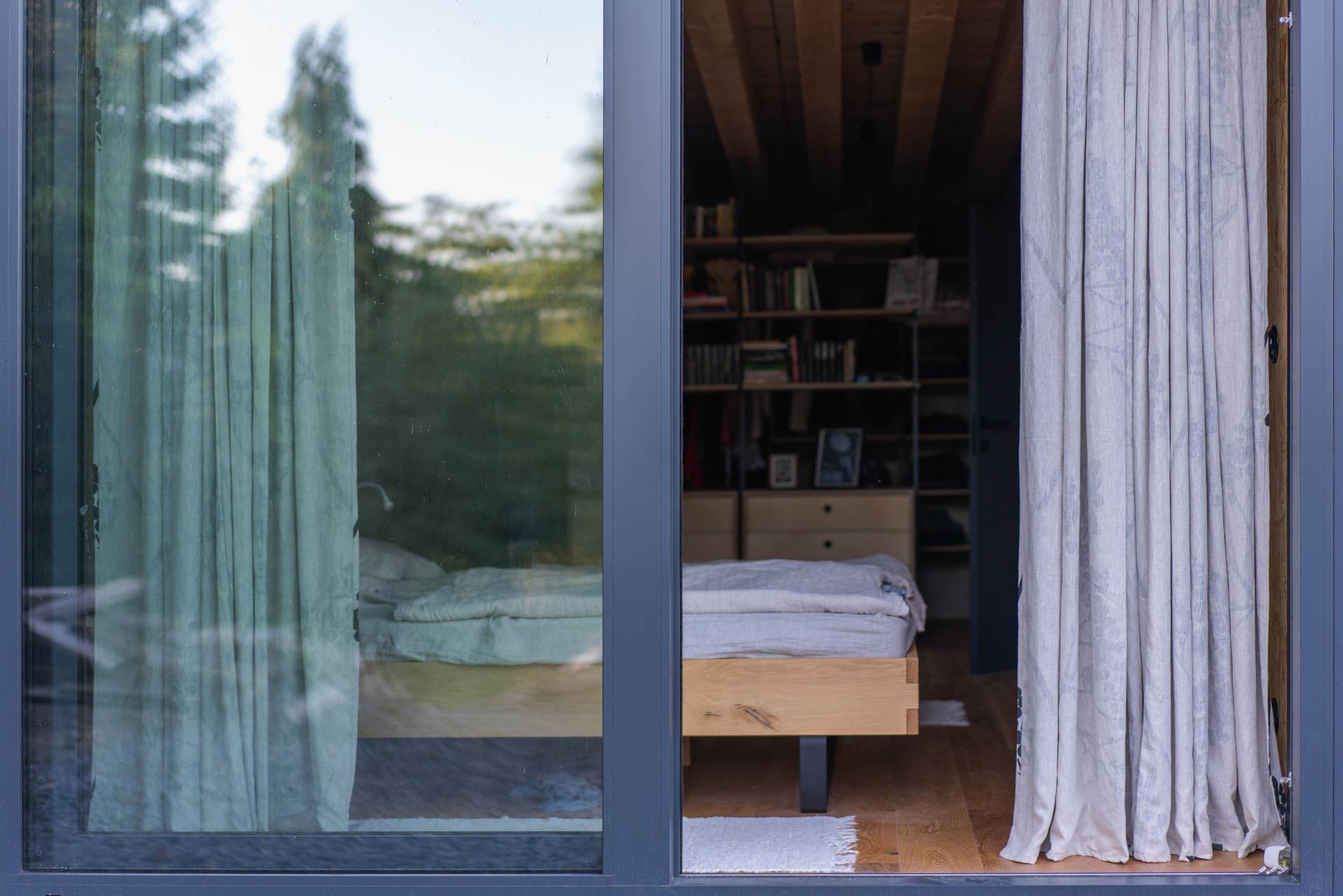 View through a glazed wall into a bedroom of the earth-sheltered house in Banská Štiavnica, wood-plank ceiling and shelves of books beyond a sliding door