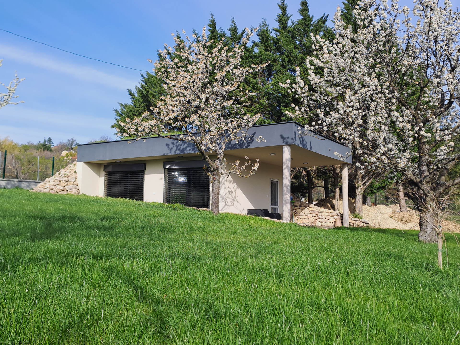 Flat-roofed earth-sheltered house with pale facade and shuttered windows, covered terrace, near Lake Balaton vineyards.