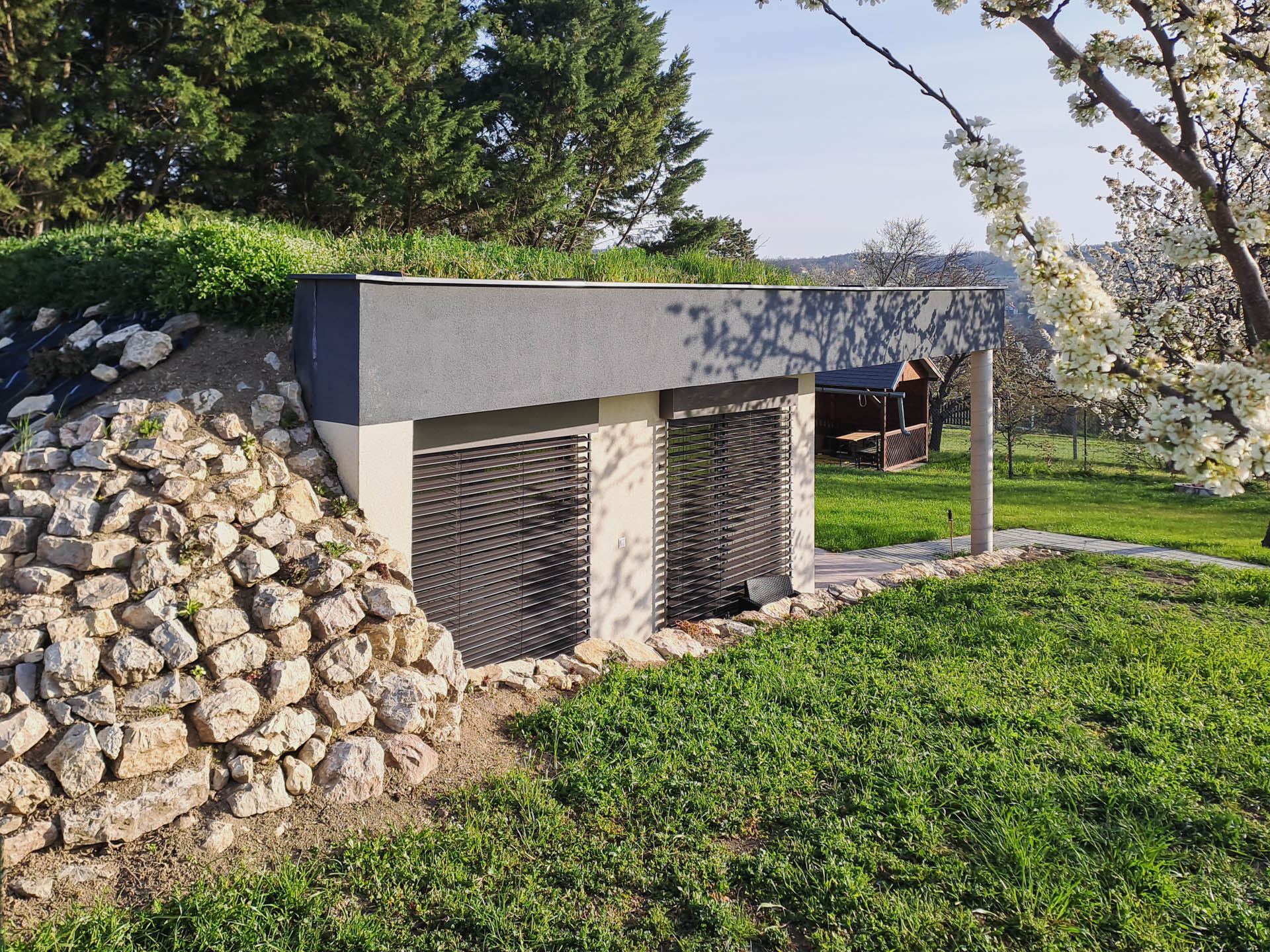 Grass-roofed earth-sheltered house at Balaton with slatted shutters, stone retaining wall and a paved walkway under a round column.