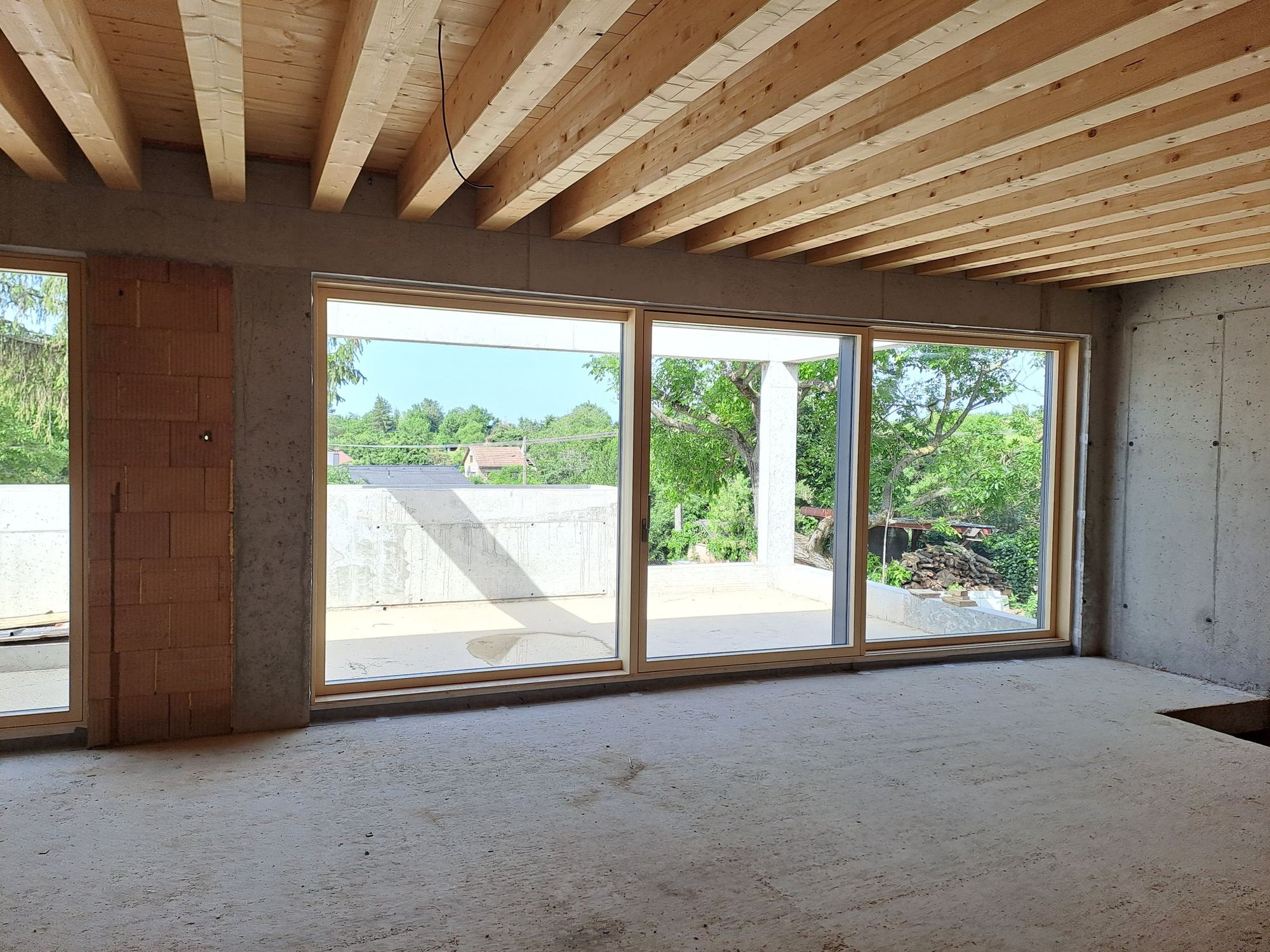 Interno grezzo di una casa interrata a Trnava, pareti in cemento e mattoni, soffitto a travi, ampia apertura verso la terrazza.
