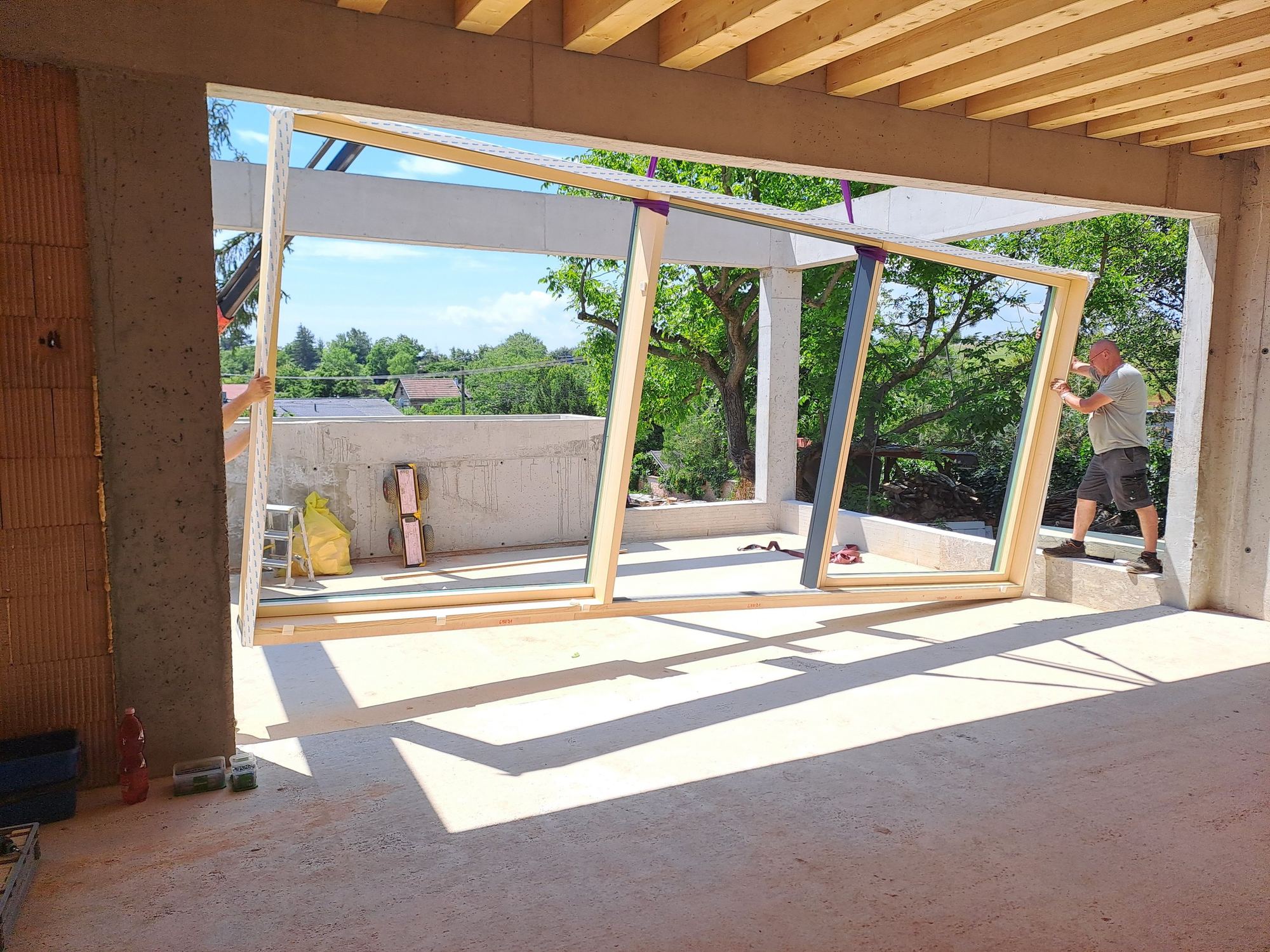 Operai inseriscono un grande infisso in un'apertura di cemento nella casa interrata in costruzione a Trnava, travi in legno a vista sopra.