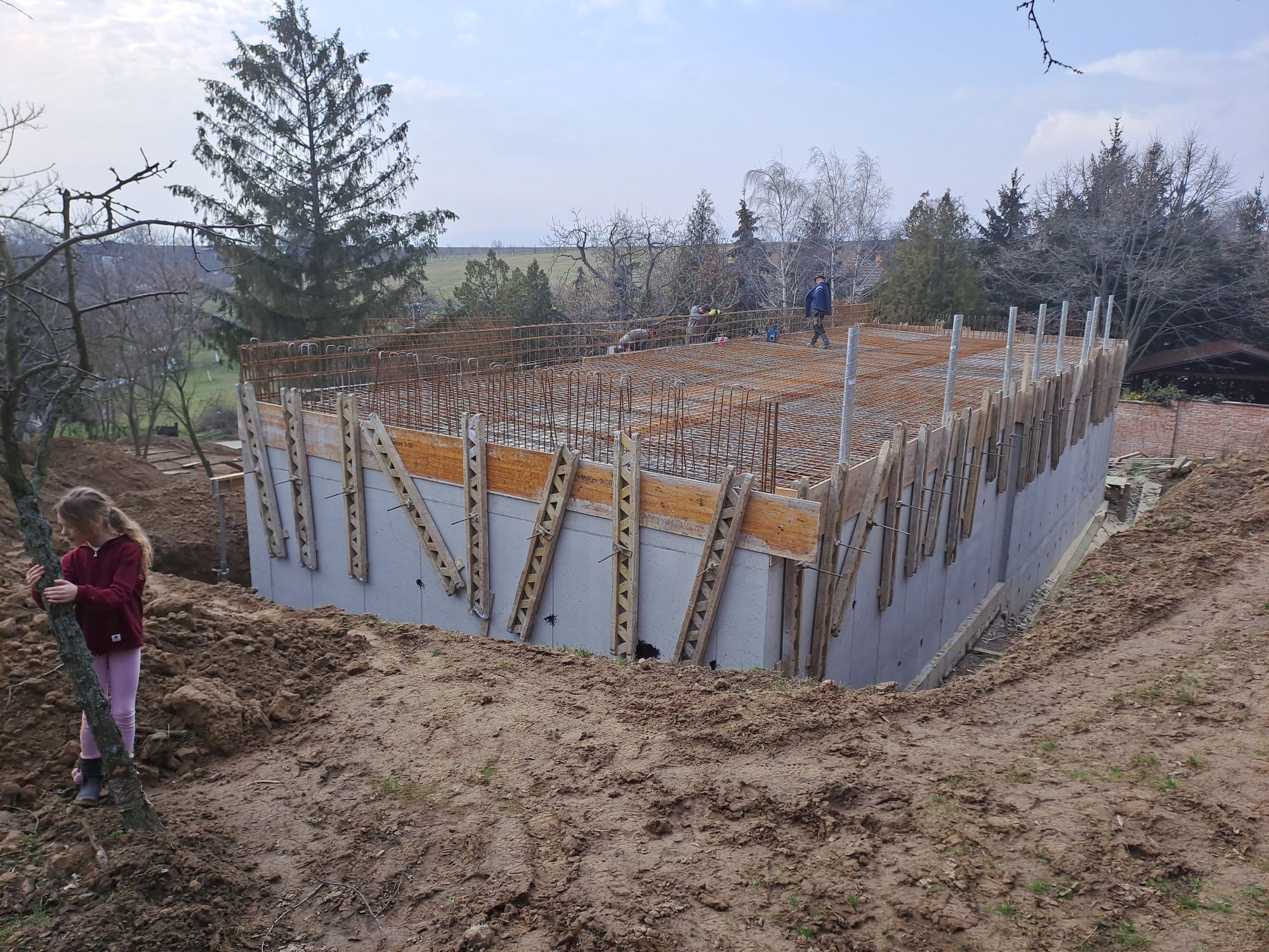 Cantiere della casa interrata di Trnava, muro in cemento con casseforme e griglia di ferri d'armatura arancioni, due operai e un bambino.