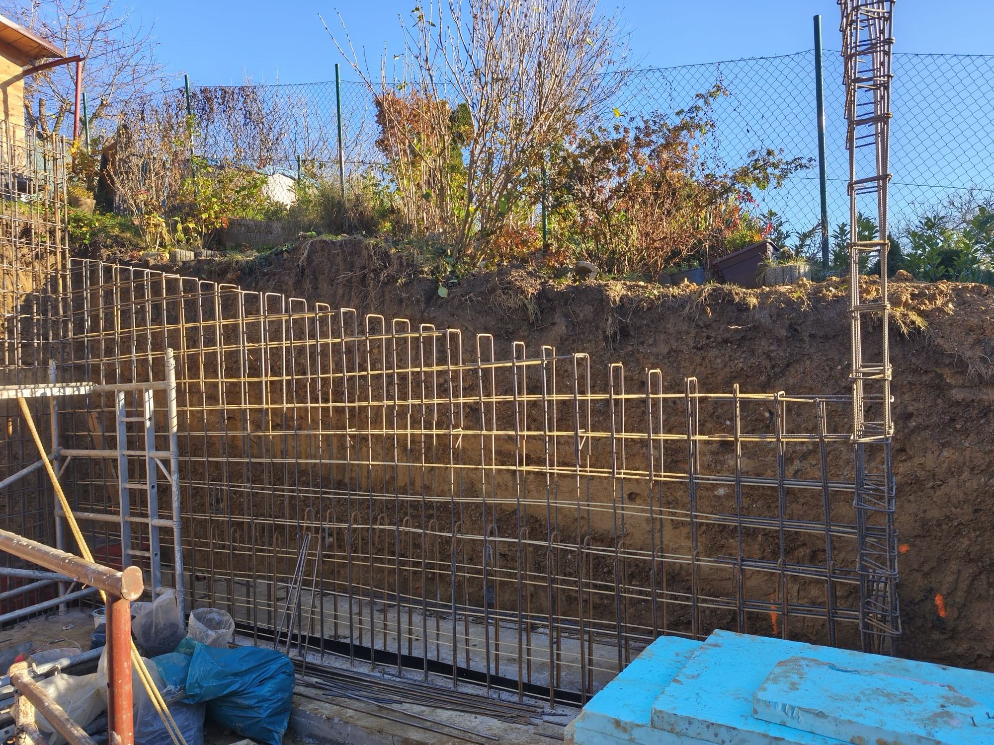 Baustelle eines Erdhauses mit Stahlbewehrung an einer Erdwand, Gerüst und blauen Dämmplatten auf der Betonplatte.