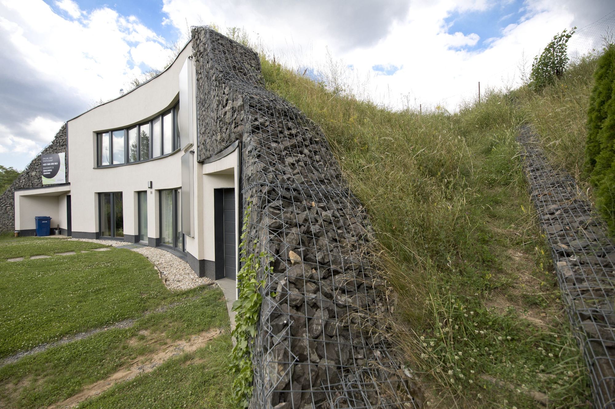 Earth-sheltered house in Rohožník with curved white facade set into a grassy slope, gabion walls flanking it