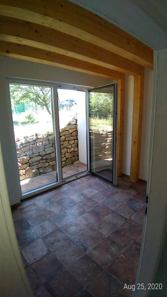 Interno di una casa interrata con soffitto curvo in travi di legno, pavimento in cotto e porta a vetri aperta su un muretto in pietra.