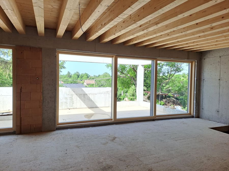 Interno grezzo di una casa interrata a Trnava, pareti in cemento e mattoni, soffitto a travi, ampia apertura verso la terrazza.