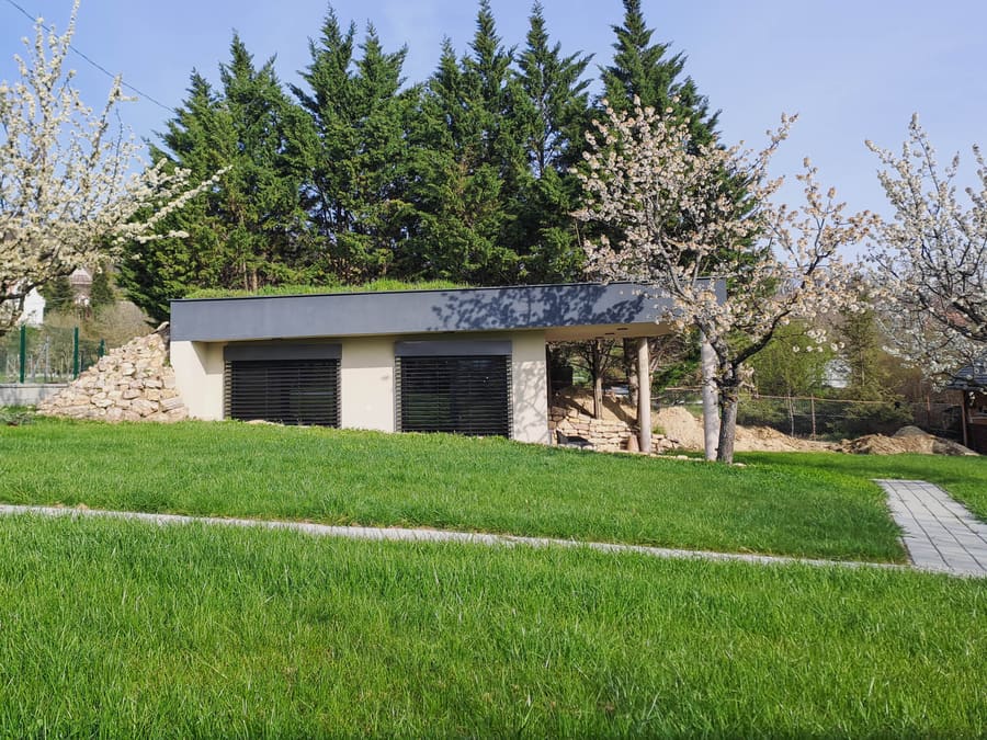 Casa interrata Balaton con tetto erboso che si fonde col prato, persiane chiuse, a destra terrazza coperta e cumulo di terra.
