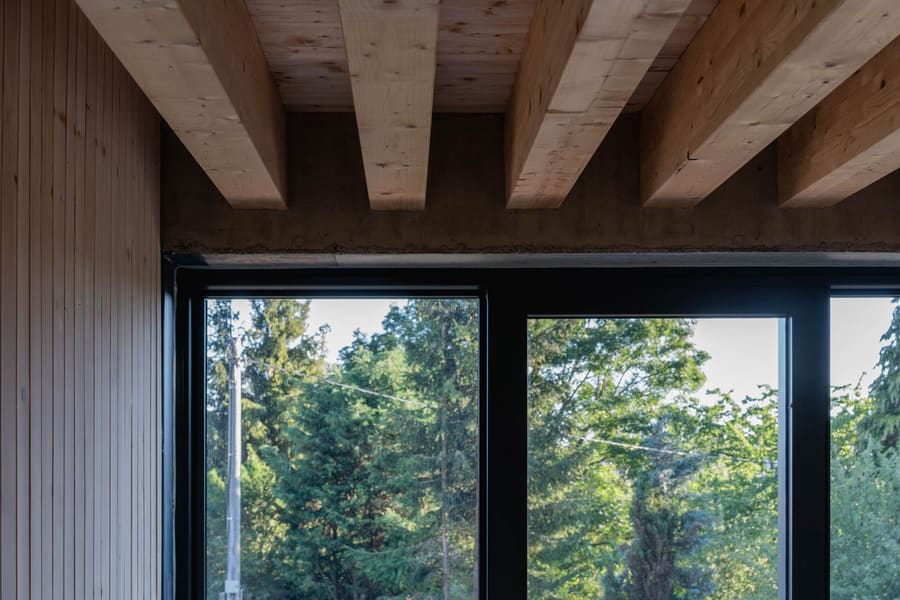 Soffitto con travi in legno e parete rivestita in legno sopra finestre dal telaio nero, alberi verdi oltre il vetro.